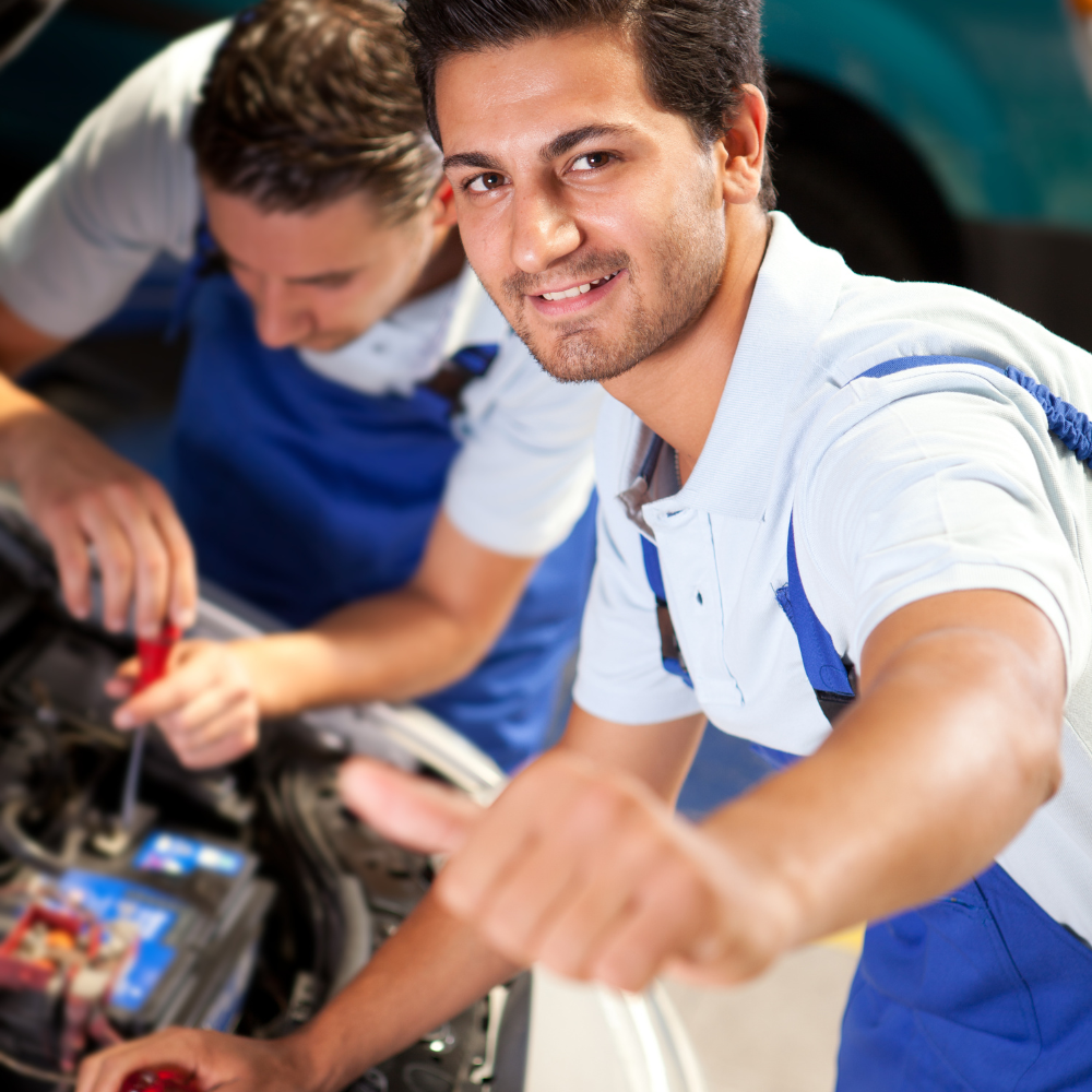 Two mechanics working on a car engine with one smiling at the camera.